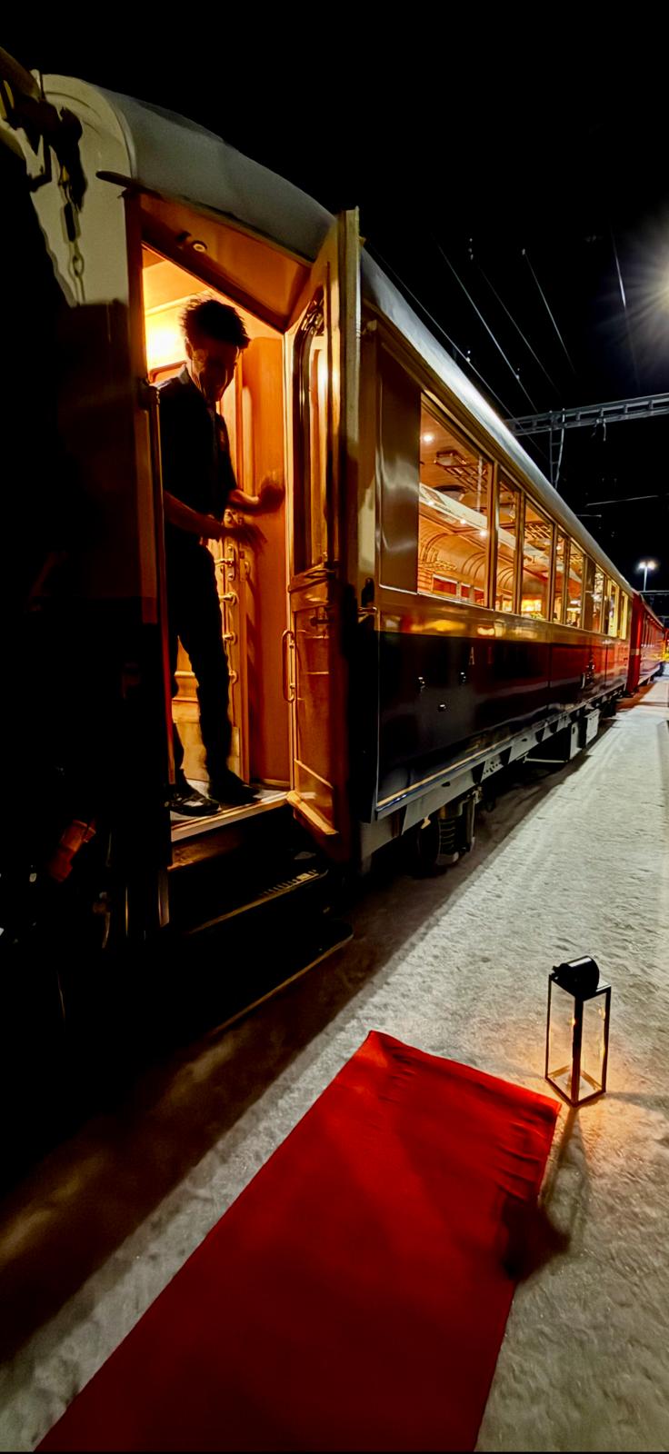 Open door of a heritage carriage at night, warm light inside, red carpet and a lantern on the snow